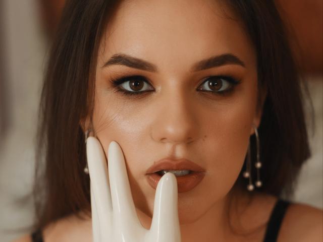 Dreia's face with brown eyes, full lips, and dark brown hair, touching her face with white, glossy fingers. She's wearing black earrings and has a slightly open mouth.