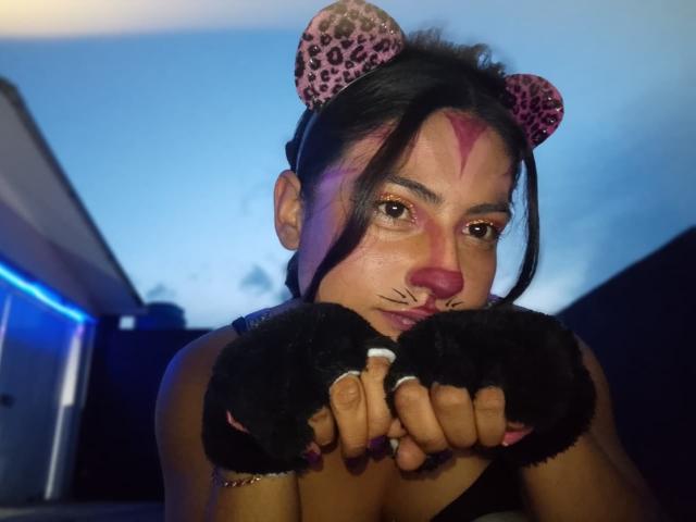 AngelAlba with pink leopard ears, black fuzzy hands, nose piercing, and ring, looking at camera with a serious expression, dark hair, pink face markings, in dim blue-lit room.