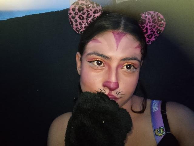 AngelAlba with leopard-print cat ears, pink face paint, black eyebrows, and holding a black plush toy against a dark background. She has a purple bra strap visible.