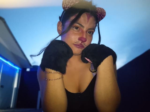 AngelAlba with leopard print cat ears, black gloves, and pink nose makeup, showing off her tits, hands up, wearing a black top, in a blue-lit room.
