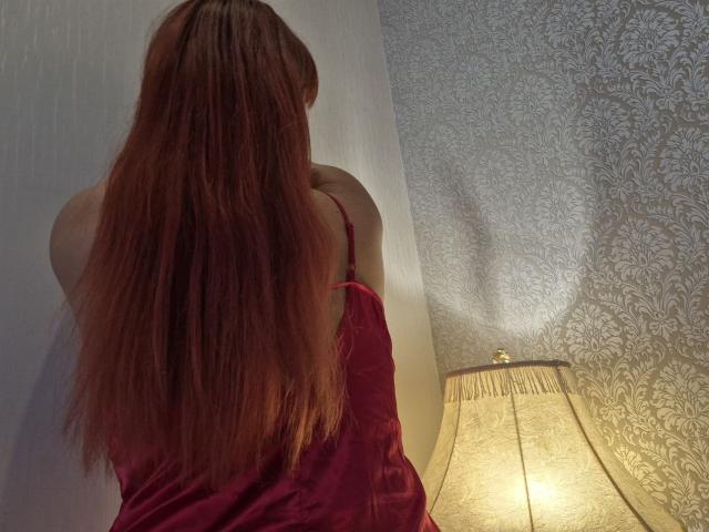 Dinastiia with long red hair, facing away, wearing a red velvet dress, standing next to a lit, patterned lampshade. Her bare shoulders are visible.