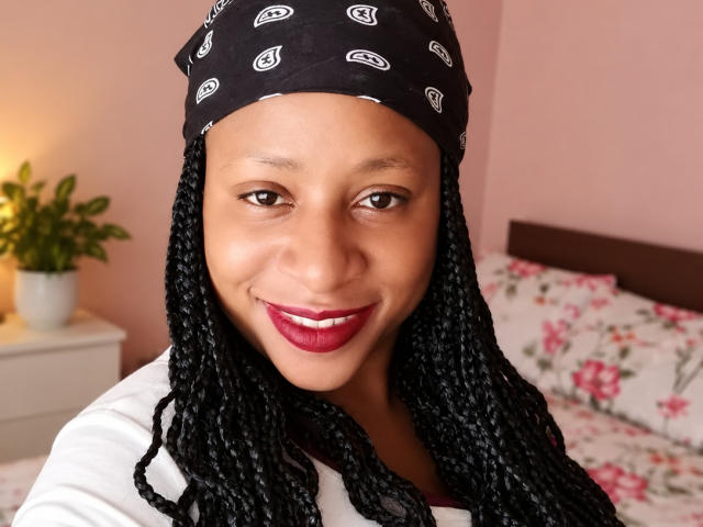 DinaRobyn, con piel oscura, largas trenzas negras, un pañuelo negro, una blusa blanca y lápiz labial rojo, sonriendo a la cámara. Sus pechos son pequeños y es muy atractiva..
