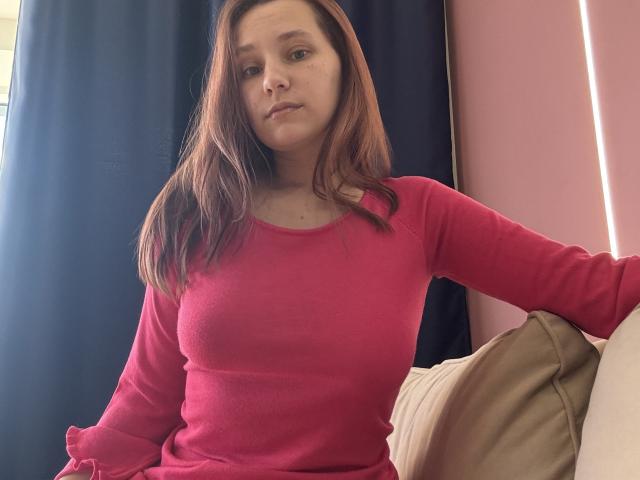 HotCaramelsTrio with medium tits, in a pink long-sleeve shirt, has brown hair, sits on a beige couch, arms resting, looking slightly bored, in a room with dark curtains.