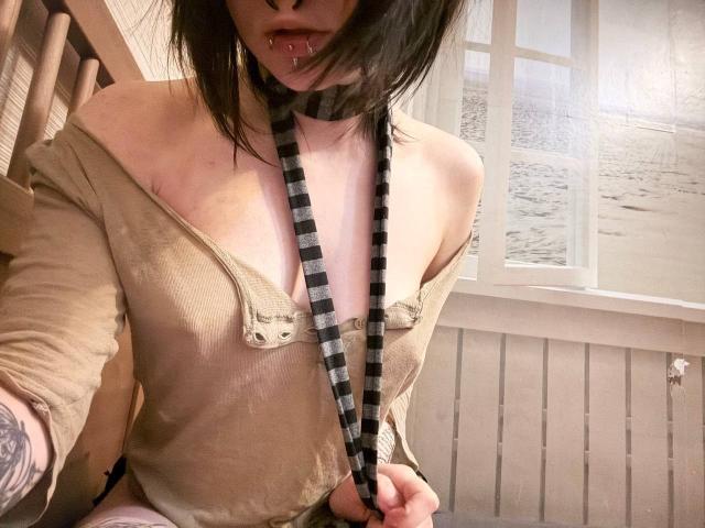 TempleWydner, avec sa peau pâle, ses cheveux noirs et son piercing au nez, porte un haut beige ample qui laisse entrevoir de petites seins, une cravate rayée noir et blanc, et un tatouage visible sur son bras..