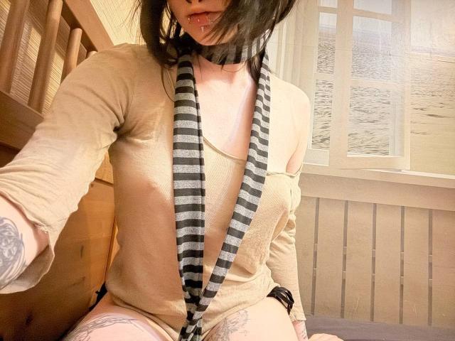 TempleWydner, avec un foulard à rayures noires, un haut beige à une épaule, et un tatouage visible sur la cuisse, montrant les mamelons, assise sur un lit en bois, la bouche légèrement ouverte, les cheveux foncés couvrant une partie du visage..