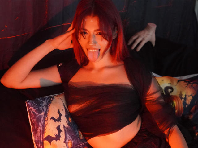 LunaBlair with red hair, black sheer top, tongue out, sitting on a Halloween-themed pillow, hand in her hair, dim red light, pale skin, visible small breasts, confident expression.