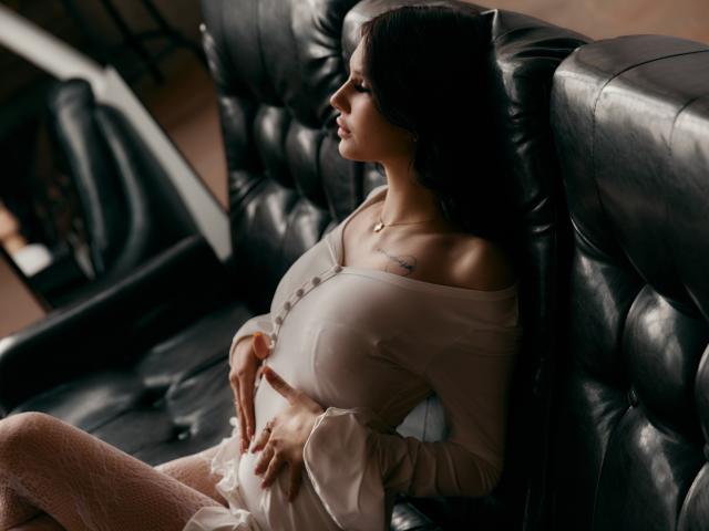 AlicePertim with large tits, wearing a beige off-shoulder top and white dress, sitting on a black leather couch, hands on her belly, dark hair, tattoo on her chest.