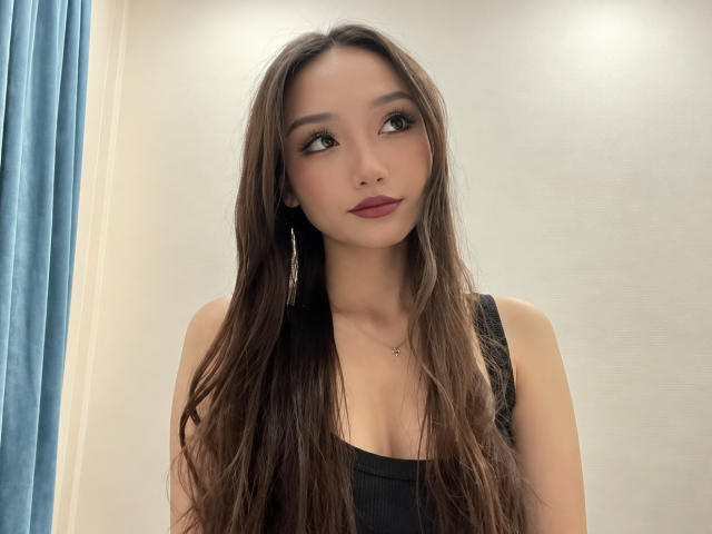 ShontaPasserino with long brown hair, wearing a black top, looking up with a slight smile, having small tits and a cute face, standing in a softly lit room.