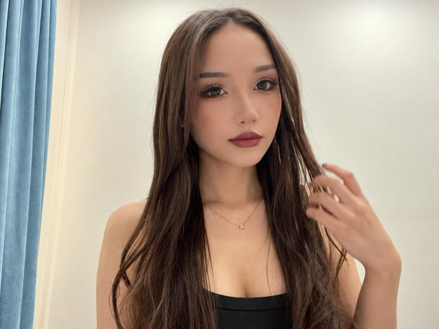 ShontaPasserino with long brown hair, dark eyes, and red lipstick, wearing a black strapless top, holding hair near her face, showing her small tits and a delicate necklace.