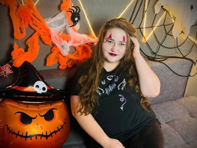 DaniellaGraceZZ con cabello castaño largo, maquillaje rojo, camisa negra y pantalones negros, sentada en un sofá gris, con un cubo de calabaza de Halloween con un sombrero de calavera, decoraciones de arañas y una red naranja..