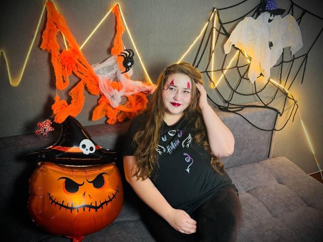 DaniellaGraceZZ with long brown hair, pink face paint, and black shirt, sitting on gray couch, next to orange jack-o'-lantern and Halloween decor, wearing a black witch hat.