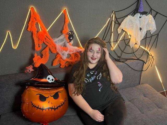DaniellaGraceZZ, chubby with long brown hair, wearing a black shirt, sitting on a gray couch, next to a glowing orange pumpkin and a witch hat with a skull.