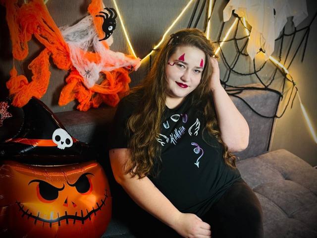 DaniellaGraceZZ con cabello castaño largo, maquillaje rojo, camiseta negra, sentada en un sofá gris, con la mano en el cabello, decoración de Halloween que incluye un cubo de calabaza, telarañas naranjas y un sombrero de bruja negro..