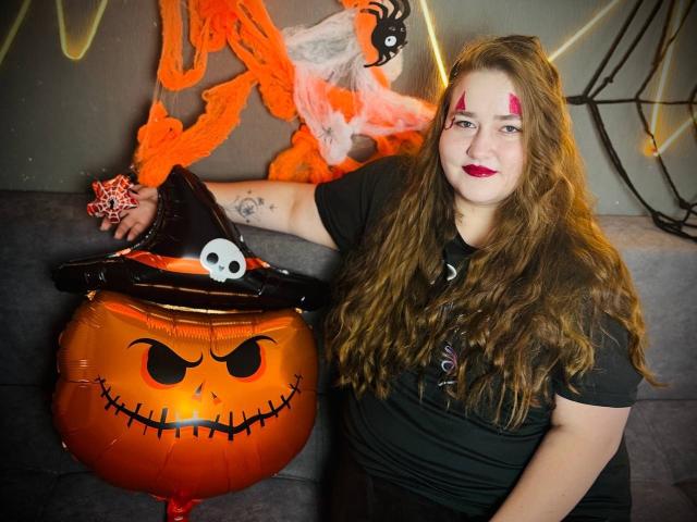 DaniellaGraceZZ with long brown hair, red makeup, black shirt, black witch hat, skull, and pumpkin balloon, standing next to a spooky orange jack-o'-lantern. Her arm tattoo visible.
