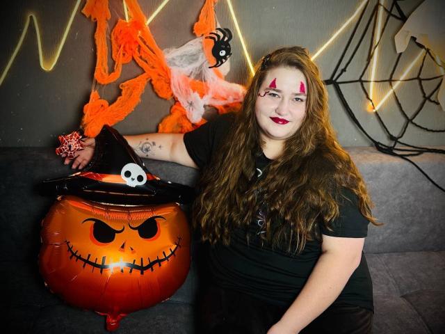 DaniellaGraceZZ with long brown hair, red face paint, black shirt, and skull hat, leaning on a red Jack-o'-lantern bucket with a grumpy face, in a Halloween setting.