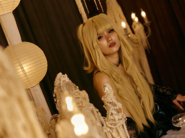 AnneTLuis with long blonde hair, blue eyes, and red lipstick, wearing a black lace top, looking over her shoulder, surrounded by lit candles and paper lanterns.