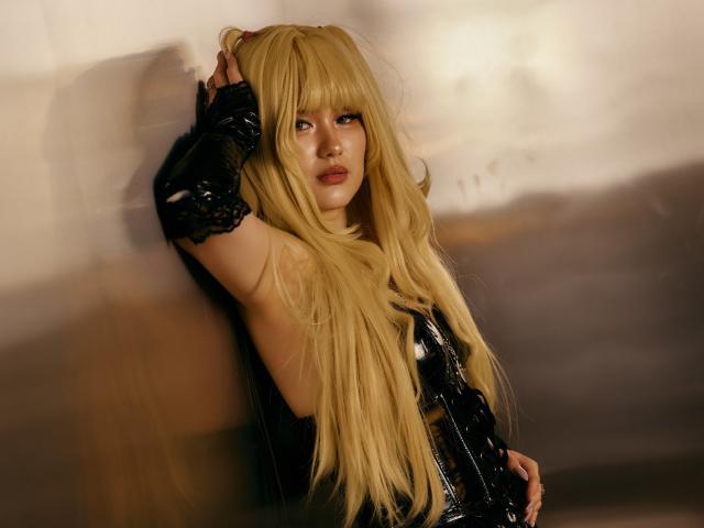 AnneTLuis with long blonde hair, black latex gloves, and black latex outfit, leaning against a wall, hand in hair, blue eyes, red lipstick, sexy, confident, and alluring.