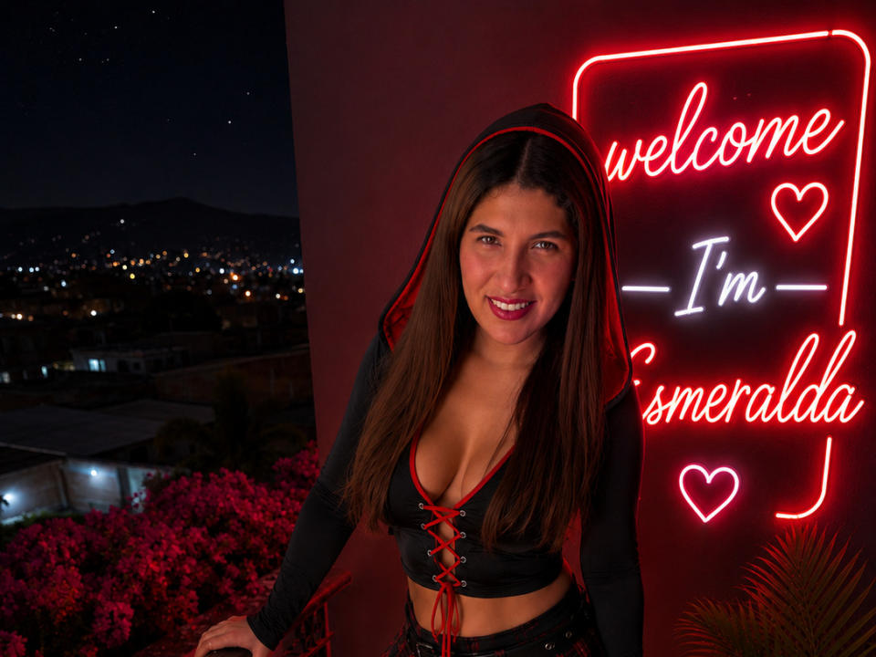 EmeraldBlonde with long brown hair, red lips, and cleavage in a black corset with red laces, smiling at a neon "Welcome I'm Smerelda" sign, night sky background.
