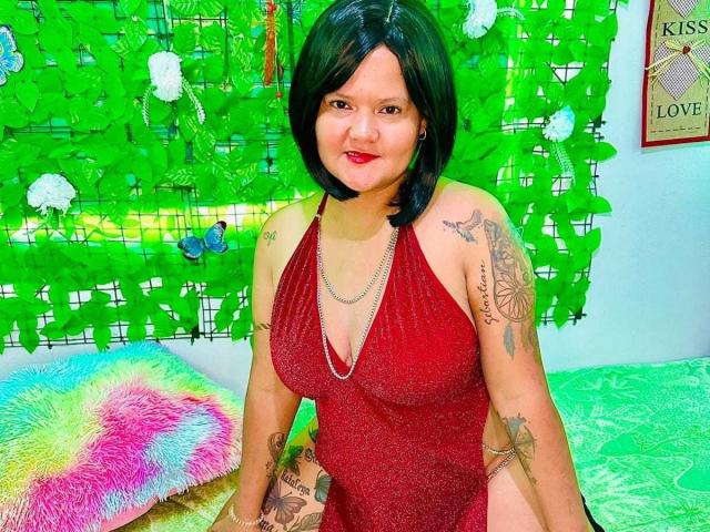 CamilaBacyc with black bob, wearing a sparkly red halter dress, showing tattoos on arms, sitting on a green bed, smiling with red lipstick, black hair, medium tits.
