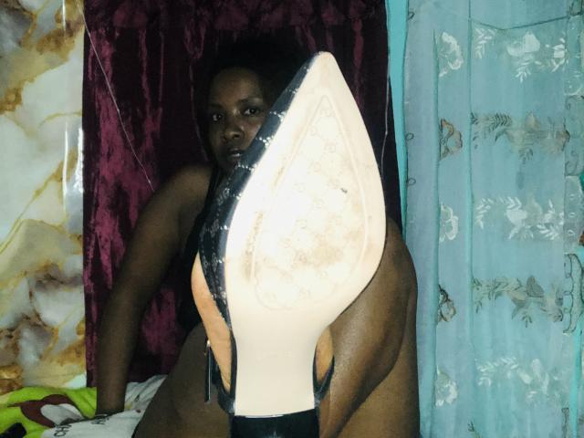TiakoI with dark skin, medium build, and small breasts, holding a dirty, white-soled high heel in front of her. She's sitting on a bed with blue and purple curtains.