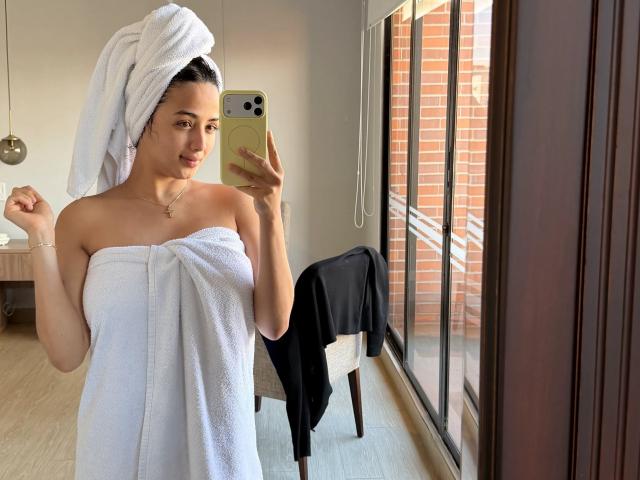 AuroraFerretti with a white towel wrapped around her ass and boobs, a towel on her head, taking a mirror selfie in a bright room with a brick window.