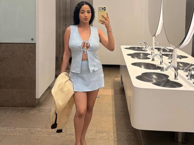 AuroraFerretti with curvy tits, wearing a light blue lace-up top and skirt, holding a beige coat, taking a mirror selfie in a bathroom with white sinks.