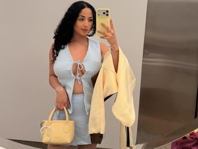 AuroraFerretti with curly black hair, light blue lace-up dress, holding yellow bag, white coat, gold phone, standing in front of mirror, smiling, wearing gold bracelet.