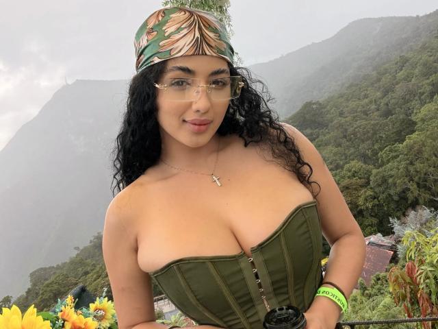 AuroraFerretti with curly black hair, green headband, glasses, and green corset, showing her big tits, wearing a cross necklace, standing in a foggy mountain landscape.