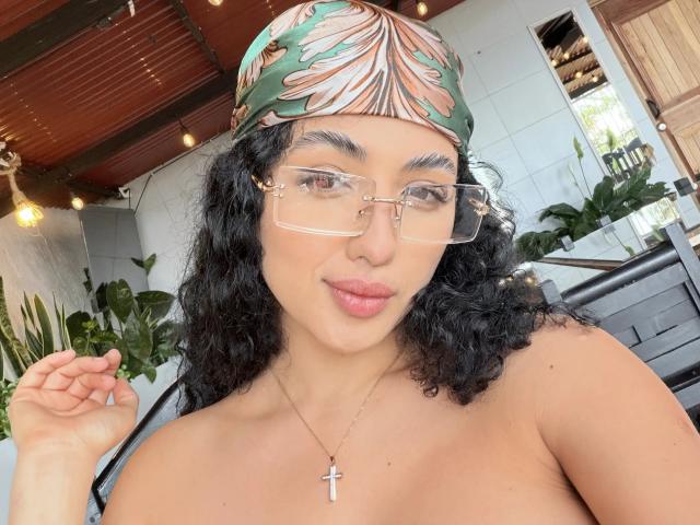 Aurora Ferretti, avec ses cheveux noirs bouclés, portant une casquette fleurie, des lunettes claires et un collier avec une croix, laissant apparaître ses épaules nues et ses lèvres roses, souriant légèrement..