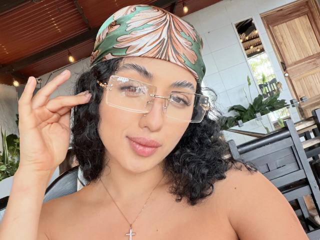 AuroraFerretti with curly black hair, green leaf-patterned headscarf, clear glasses, nose ring, cross necklace, and pink lips, holding her glasses, showing her bare shoulders.