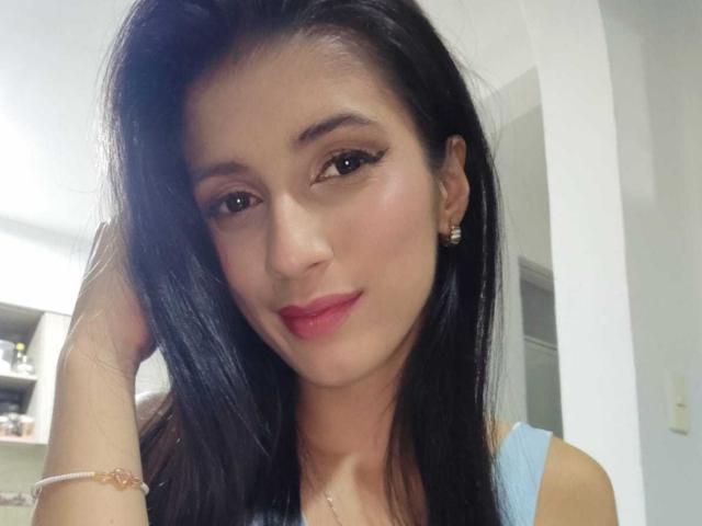 JasminKooper tem cabelo preto escuro, olhos castanhos, batom vermelho e está vestindo uma blusa azul clara com uma pulseira rosa. Ela está olhando para a câmera com um leve sorriso..