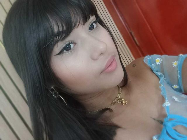 AshlyLee with dark straight hair, blue eyes, and black eyeliner, wearing a sheer blue top with daisy patterns, and a gold necklace, smiling slightly.