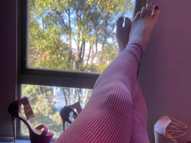 RossSofiia's legs are up, wearing pink ribbed leggings, with her feet against a window, showing her bare soles. Pink high heels and a white shoe are below.