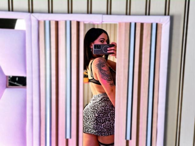 MarianaJackson with long black hair, tattooed arm, wearing a black bra and leopard print skirt, taking a mirror selfie, showing her butt and back, holding a smartphone.
