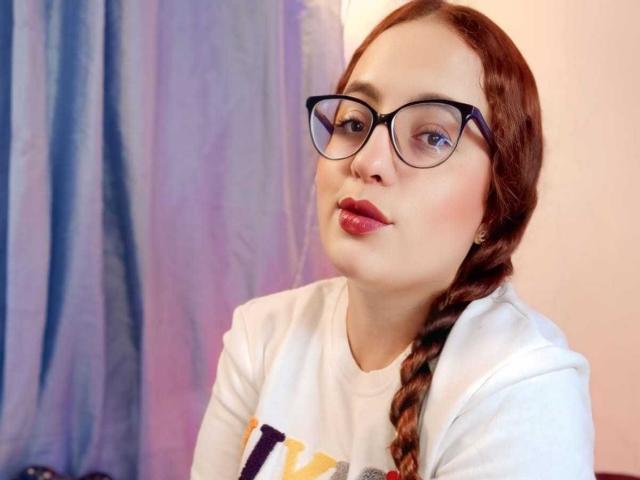 StacyMillerr with brown braided hair, black glasses, and pink lipstick, wearing a white shirt, pouting slightly with her lips. Her ass is not visible.