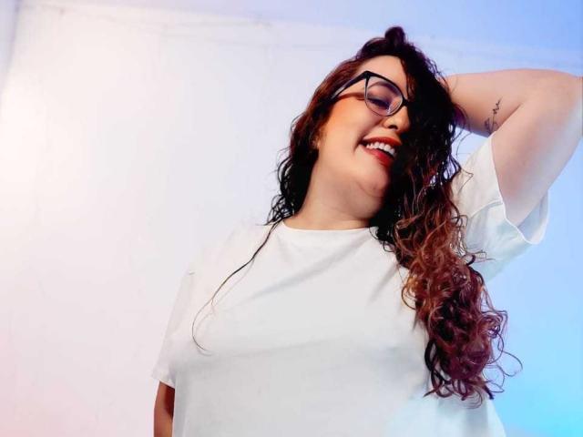 StacyMillerr with curly brown hair, glasses, and white shirt, smiling, raising arm, showing tattoo on side, has big tits and a cute face.