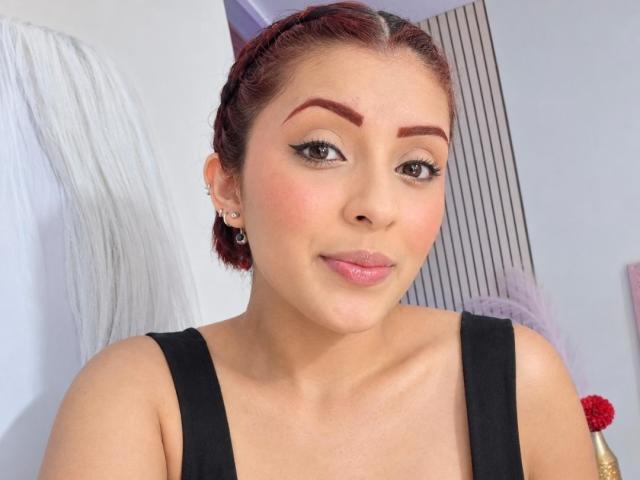 LizBonnet hat rote, geflochtene Haare, dicke Augenbrauen, Eyeliner im "Cat-Eye"-Stil, pinken Lippenstift und trägt ein schwarzes Oberteil, das ihr süßes Gesicht und ihr kleines Hinterteil zeigt. Sie lächelt sanft..
