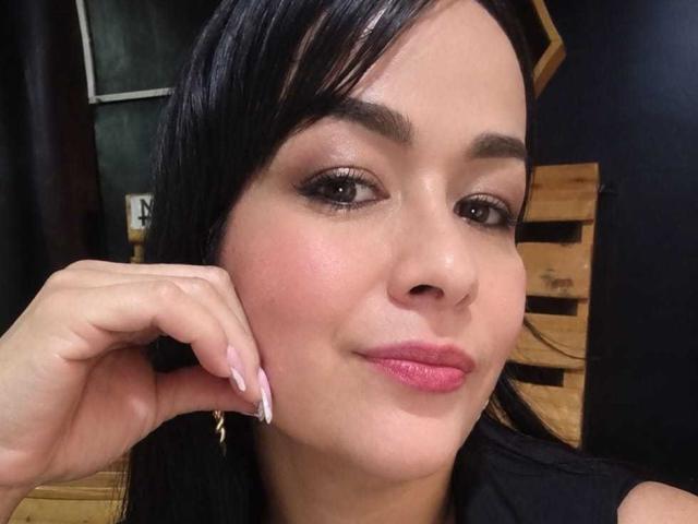 TamaraFoxy hat dunkles Haar, braune Augen und rosafarbenen Lippenstift, mit einem leichten Rouge auf ihren Wangen. Sie legt ihre Hand auf ihr Gesicht und zeigt ihre langen, rosafarben lackierten Finger..