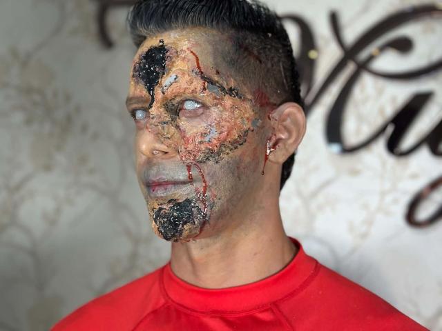 O rosto de JoseRumBerotop está coberto por uma mistura de tinta preta e vermelha, com um pouco de sangue seco, cobrindo seu nariz, bochecha e testa. Ele está vestindo uma camisa vermelha e olhando para o lado..