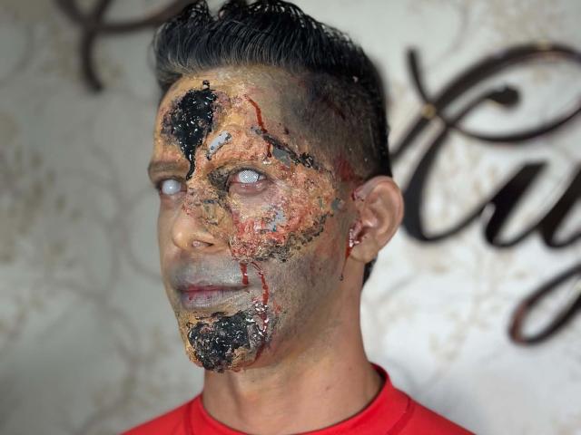 JoseRumBerotop, con un trucco opaco e screpolato sul viso, capelli corti e scuri, occhi azzurri pallidi, una camicia rossa e un piccolo orecchino, appare serio con un aspetto sporco e ruvido..