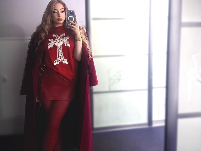 ElviraCrichlow with long blonde hair, red dress, and coat, holding phone, wearing a large white cross necklace, looking in mirror, showing her ass, and red tights.