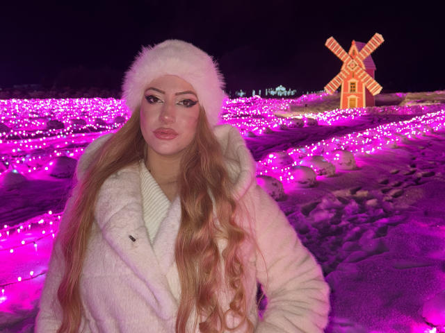 ElviraCrichlow with long blonde hair, white furry hat, and white coat, standing in a pink-lit snowy field, has pink-illuminated eyes, full pink lips, and a sexy, cute face.