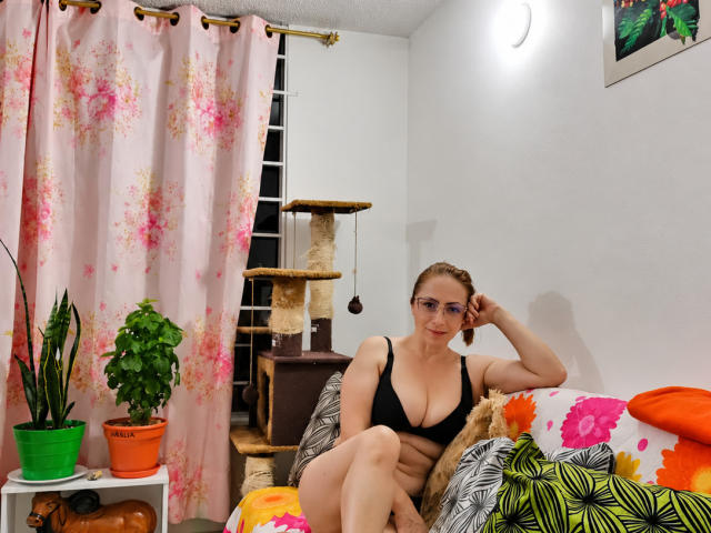GianineLarose, wearing a black bra, sits on a chair with a cat on her lap, showcasing her large boobs and pale skin, with potted plants and a cat tree.