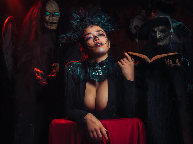 Barbara Leythonn, avec de grandes seins, des cheveux noirs et un maquillage gothique, portant un collier noir, un haut en cuir et assise à une table rouge, tenant un livre, entourée de masques de crânes effrayants..
