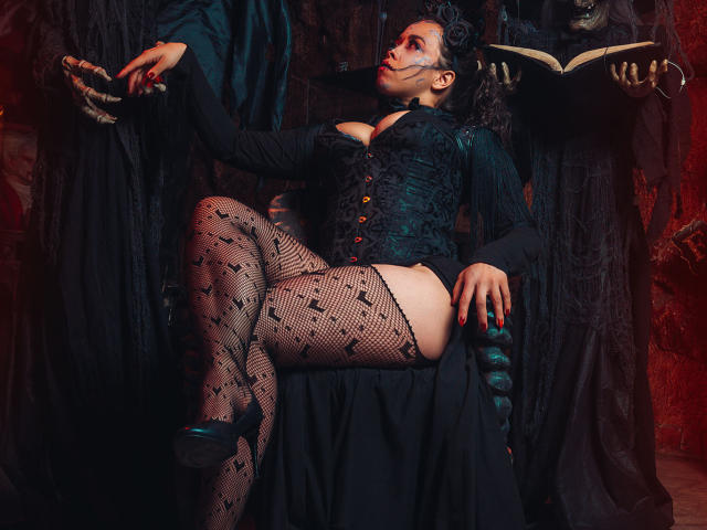 Barbara Leythonn, vêtue de manière gothique, est assise avec une jambe levée, exposant ses collants résille, son corset noir à lacets et ses fesses nues. Elle tient un livre ouvert, avec un maquillage sombre..