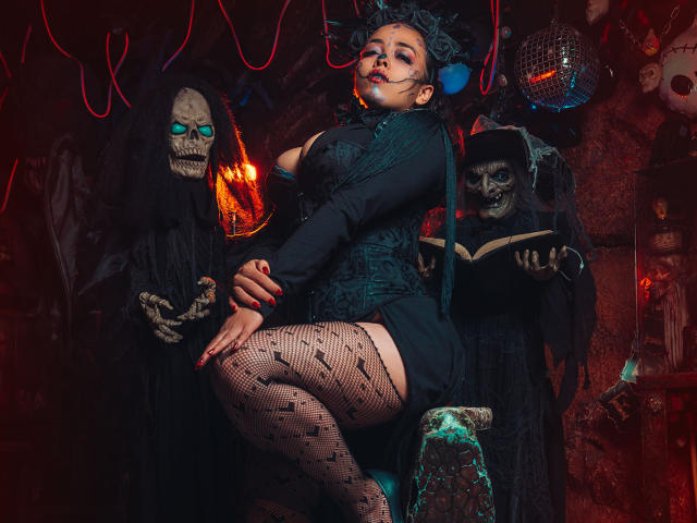 Barbara Leythonn, con calze a rete scure e decorate, un abito nero con lacci e un copricapo blu, è affiancata da figure con maschere a forma di teschio. Sullo sfondo, luci al neon rosse, una stanza buia e una palla da discoteca..