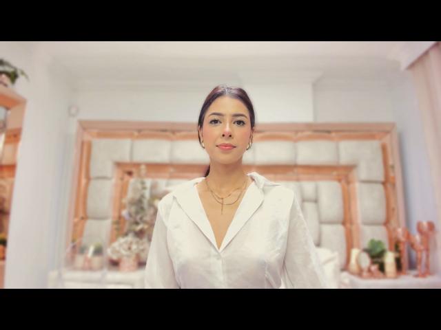 HellenBells has dark brown hair, medium tits, wearing a white shirt, standing in a room with a wooden headboard, and a gold necklace, looking straight at the camera.
