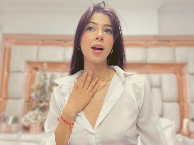 HellenBells has dark brown hair, a white shirt, and a red bracelet. She's touching her neck with her hand, mouth slightly open, looking upward. Her tits are visible.