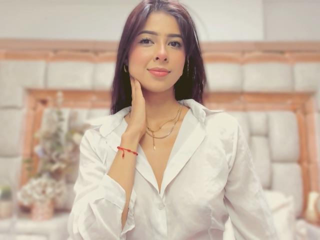 HellenBells with long dark hair, wearing a white button-up shirt, smiling, touching her cheek, gold necklace, red bracelet, soft light, bedroom, looking cute and sexy.