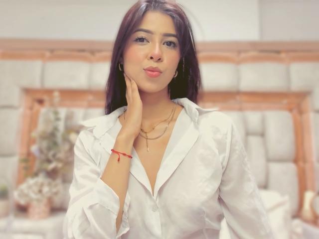 HellenBells with dark hair, pouty lips, wearing a white button-up shirt, hand on cheek, gold necklace, red bracelet, looking at camera. Boobs slightly visible through shirt.